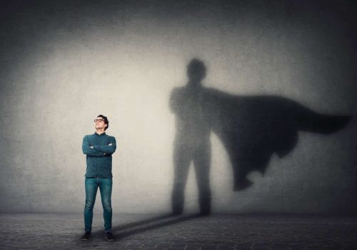 Brave man keeps arms crossed, looks confident, casting a superhero with cape shadow on the wall. Ambition and business success concept. Leadership hero power, motivation and inner strength symbol.