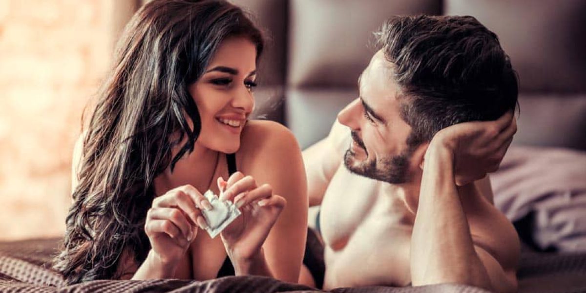 Sexy young couple is looking at each other and smiling while lying on bed at home, woman is holding a condom